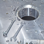 Zwei offene Maulschlüssel liegen auf einer großen, runden Metallplatte mit vielen kleinen Bohrungen und einer zentralen Öffnung. Die Oberfläche wirkt bearbeitet und industriell, vermutlich Teil einer größeren mechanischen oder technischen Konstruktion.