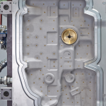 Ein präzise bearbeitetes Metallbauteil mit vielen kleinen, regelmäßig angeordneten Bohrungen und einer zentralen, goldfarbenen, runden Einlassung. Das Teil ist von einem umlaufenden, metallisch glänzenden Rand eingefasst und scheint Teil eines technischen oder industriellen Systems zu sein, möglicherweise ein Werkzeug- oder Formeinsatz.