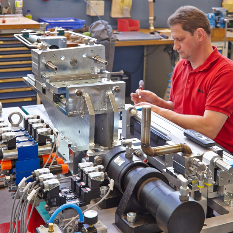 Ein Techniker arbeitet an einem Prüfstand mit hydraulischen und elektronischen Bauteilen in einer Werkstatt.